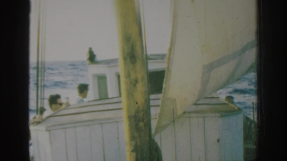 1962 Rhodes travel footage showing a white sailboat with a weathered pale sail, mast, rigging, and passengers on calm blue water.