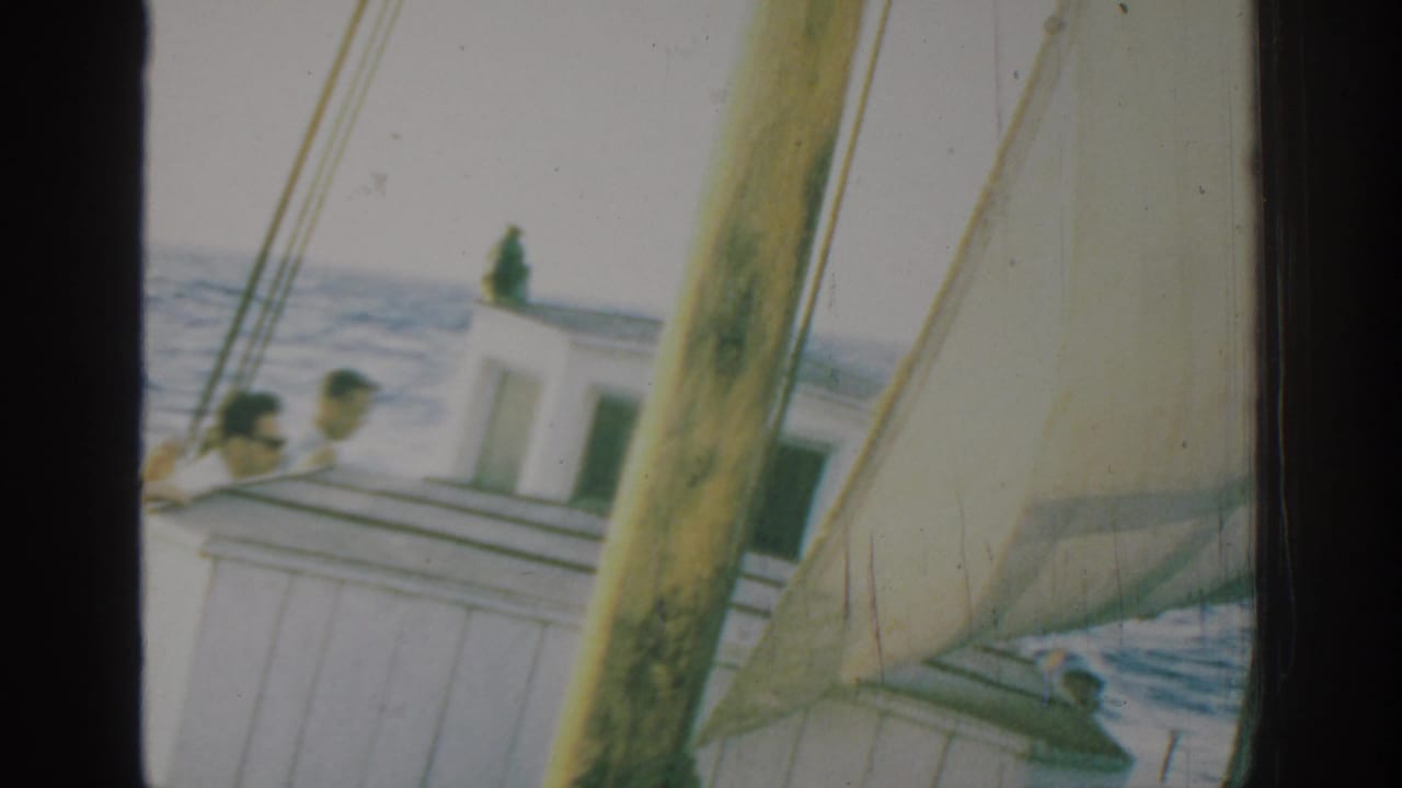 1962 Rhodes, Greece home-movie frame showing passengers on a white sailboat cabin as mast, sailcloth, rigging, and blue sea fill the view.