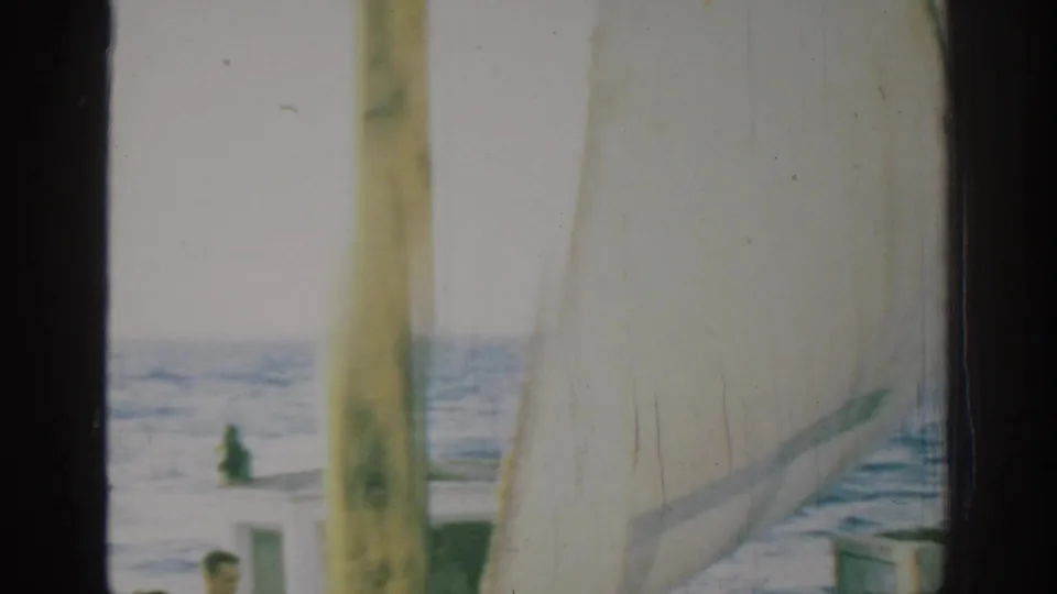 1962 Rhodes, Greece frame with a yellow mast and pale sail in the foreground, open sea beyond, and part of a passenger near the cabin.