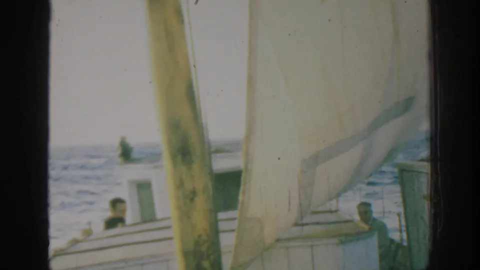 1962 Rhodes, Greece home-movie frame showing the sailboat cabin and passengers partly obscured by mast and sail, with choppy sea behind.