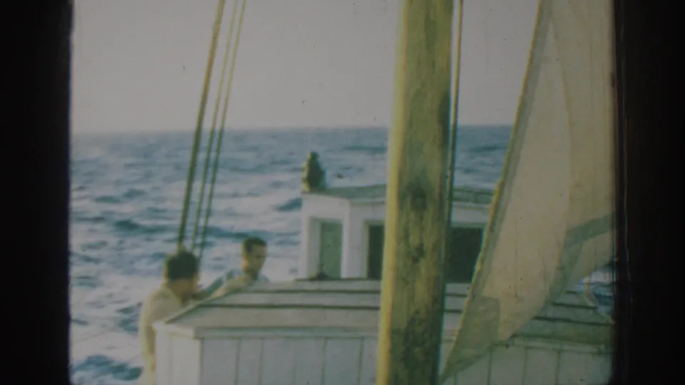 1962 Rhodes, Greece frame showing two passengers by the white cabin of a sailboat, with mast, sailcloth, rigging, and blue sea around them.