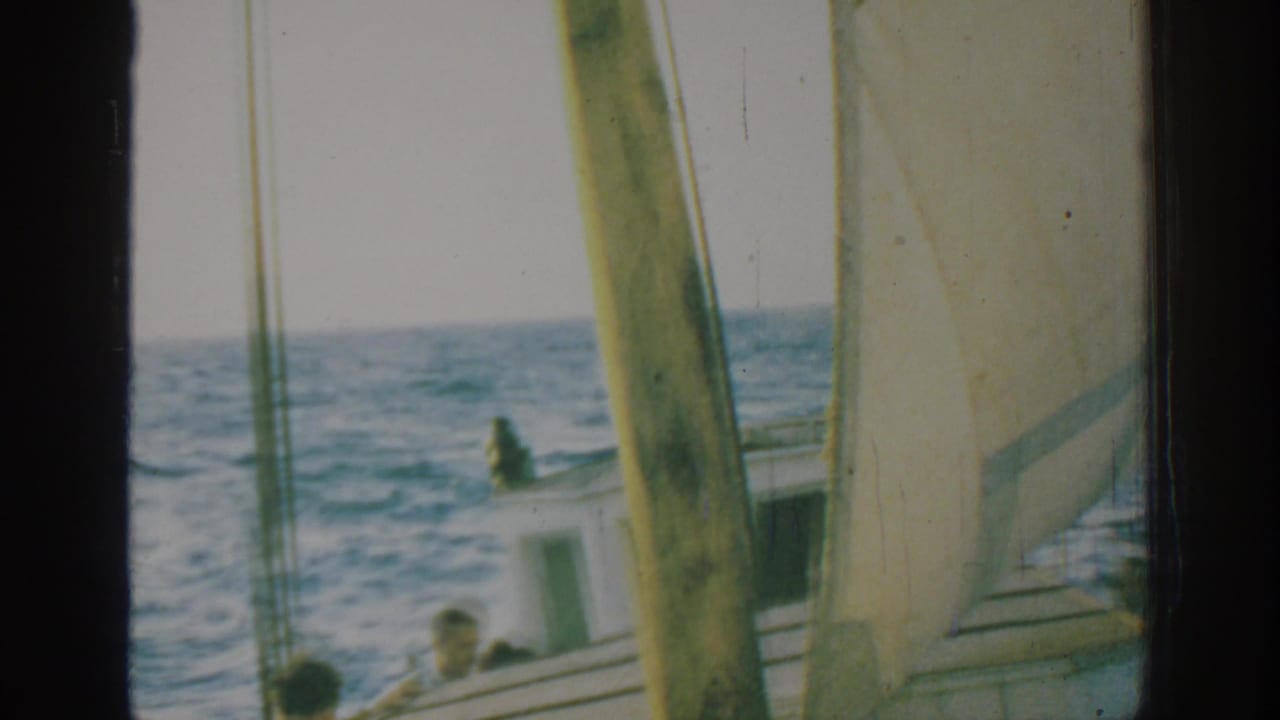 1962 Rhodes home-movie frame showing a small sailboat, white sail, mast, and men on deck amid choppy blue water.