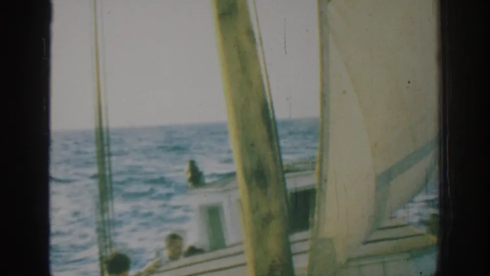 1962 Rhodes home-movie frame showing a small sailboat, white sail, mast, and men on deck amid choppy blue water.