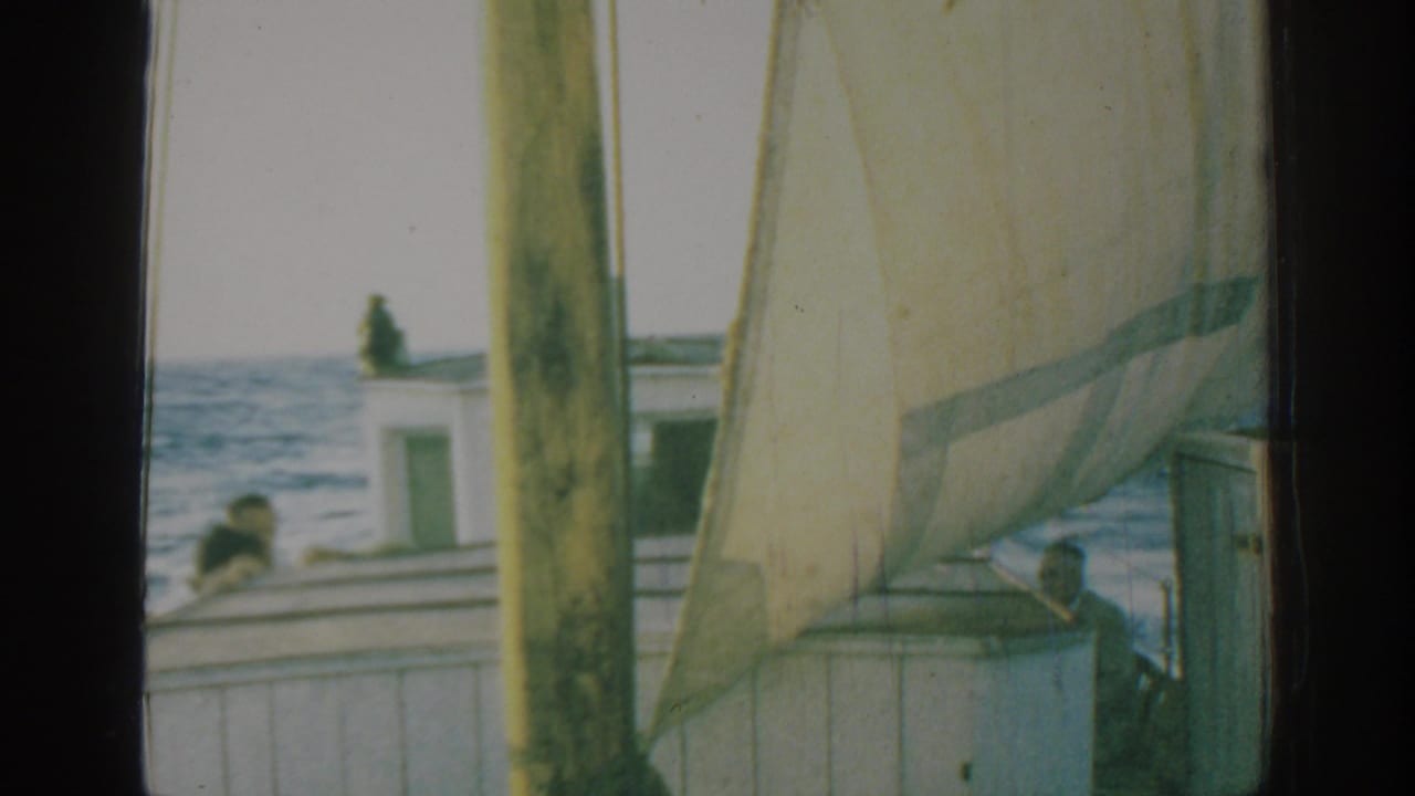 1962 Rhodes sailing frame with the sail, mast, rigging, deck cabin, and a man near the bow against moving water.