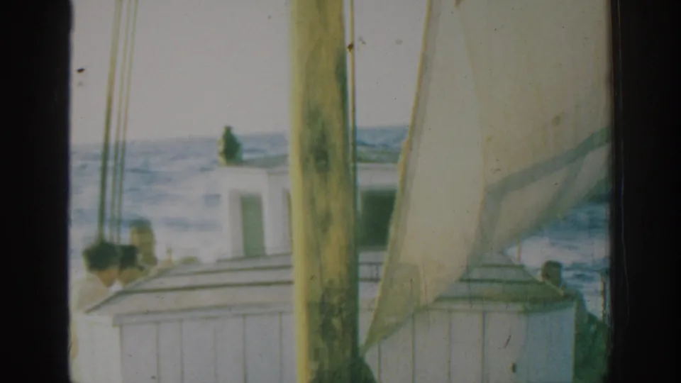 1962 Rhodes home-movie frame showing men clustered near the rail of a small sailboat with sail, mast, and choppy sea behind.