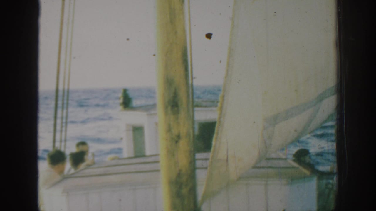 1962 Rhodes, Greece 8mm frame showing passengers on a sailboat deck beside a white cabin, with mast, pale sail, and open sea beyond.