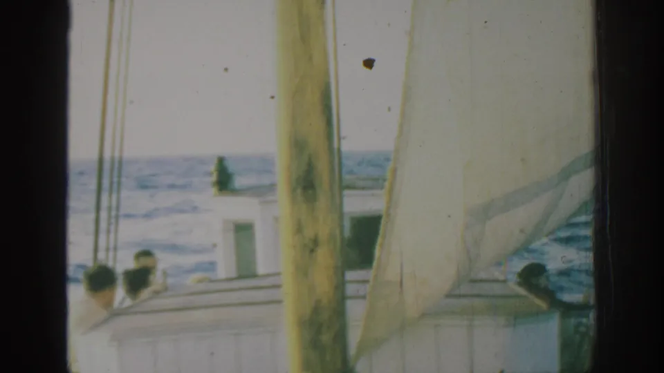 1962 Rhodes, Greece 8mm frame showing passengers on a sailboat deck beside a white cabin, with mast, pale sail, and open sea beyond.