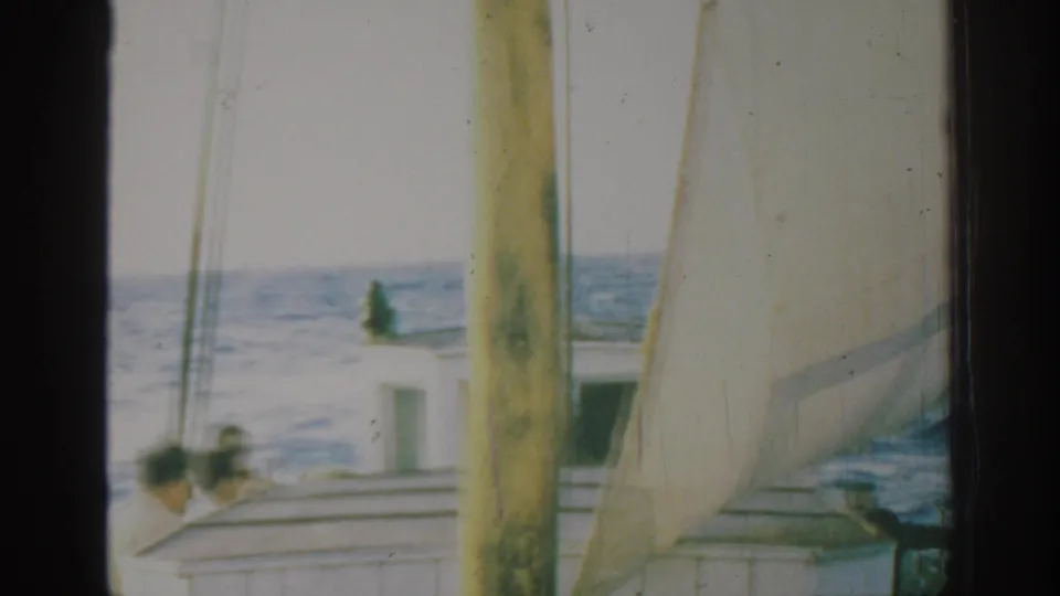 1962 Rhodes, Greece 8mm frame with a sailboat cabin and passengers partly obscured by mast, sail, rigging, and ocean horizon.
