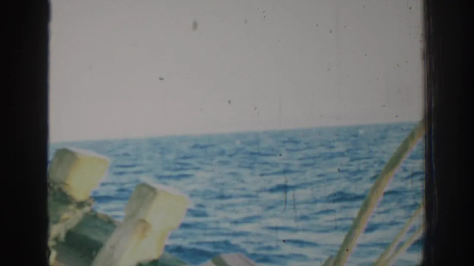 1962 Rhodes home-movie frame showing blue open water beyond weathered wooden boat posts and dark 8mm frame edges.