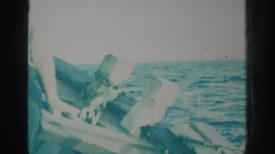 1962 Rhodes home-movie frame with a briefly visible seated person in a wooden boat, surrounded by blue open water.
