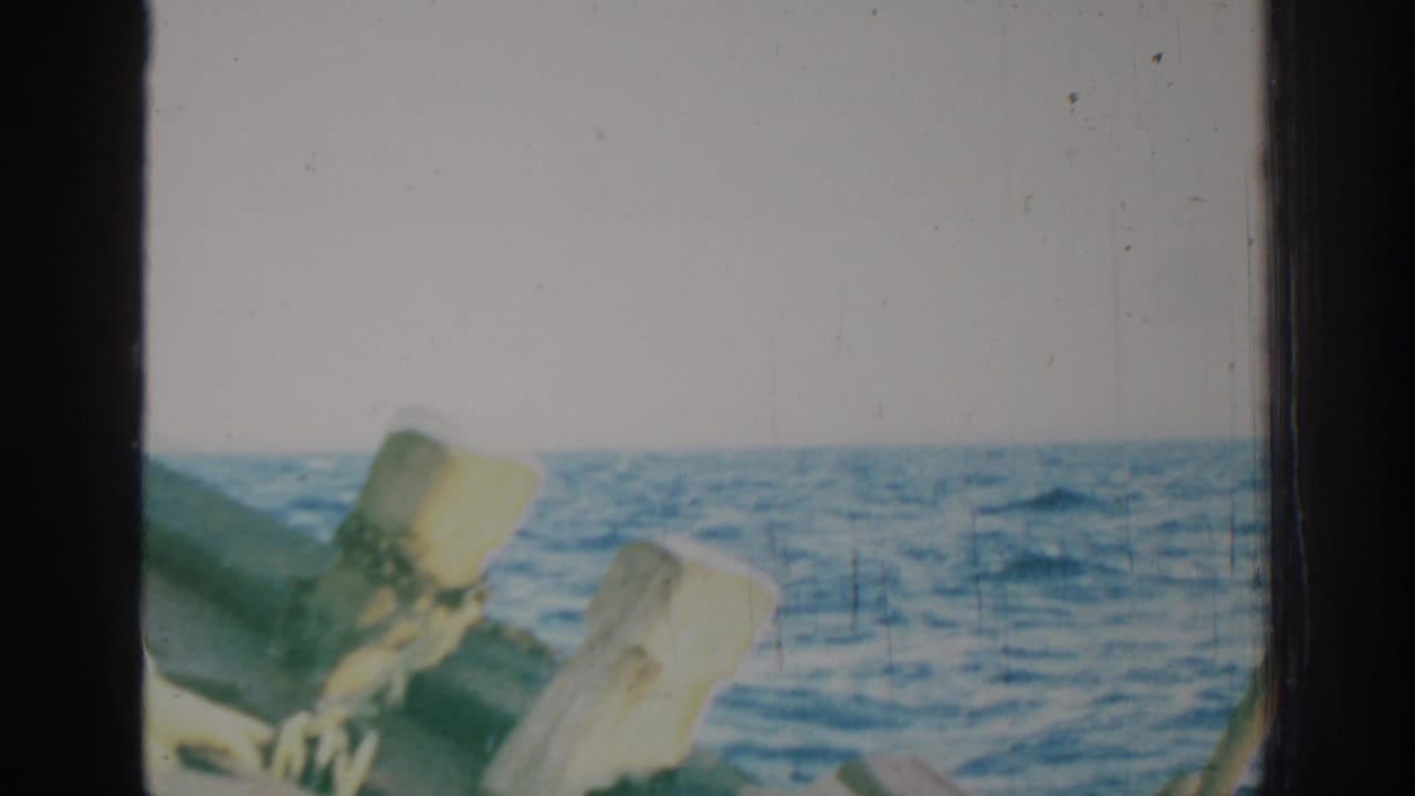 1962 Rhodes home-movie frame showing blue sea, pale sky, dark boat edge and weathered wooden posts in the foreground.