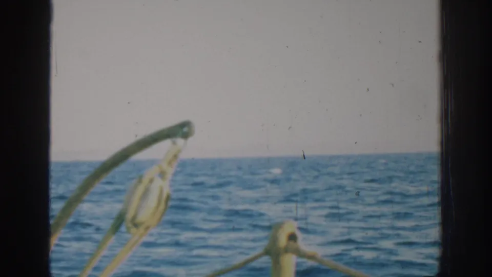 A 1962 Rhodes boat-view frame showing open water, pale sky, rigging and a curved metal fitting close to the camera.