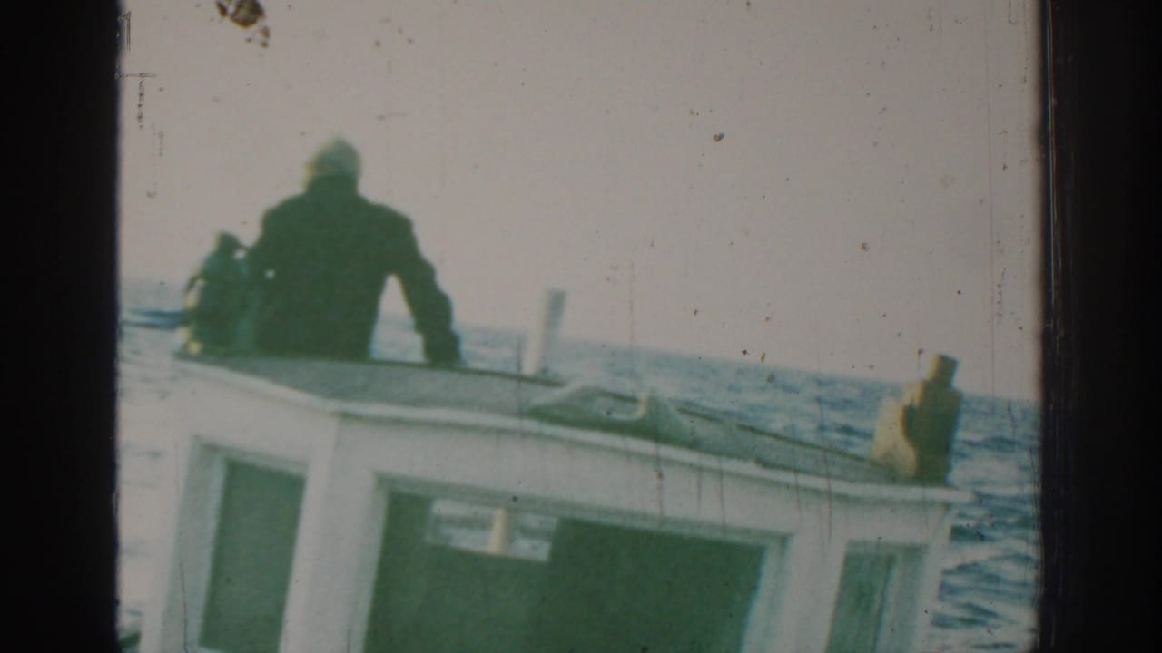 Man in dark clothing standing near the bow of a small boat on open water.
