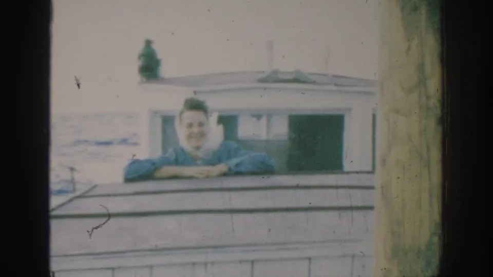 1962 Rhodes home-movie frame showing a woman in blue leaning on the cabin edge of a small boat at sea.