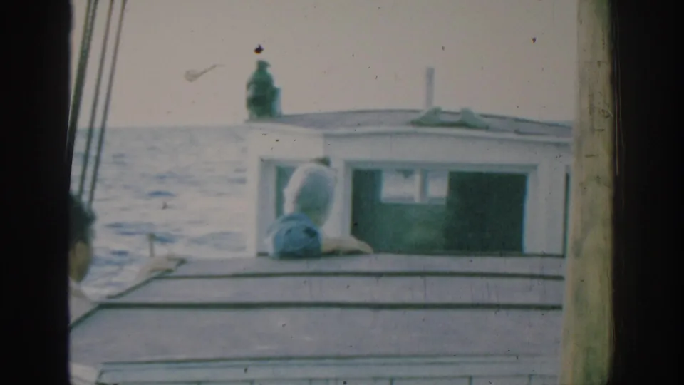 1962 Rhodes frame showing a woman in blue turned away while leaning on the cabin of a small boat on open water.