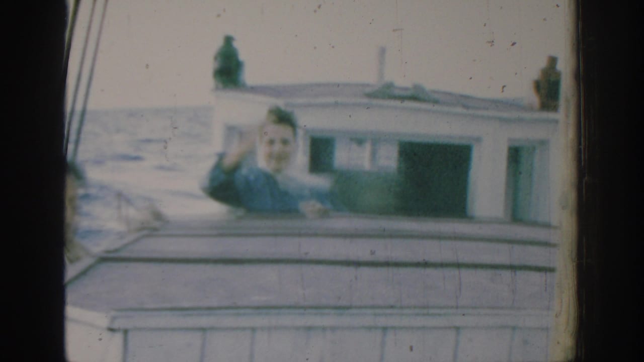 In Rhodes, Greece, 1962, a woman in a blue shirt waves toward the camera from a small boat on the sea.