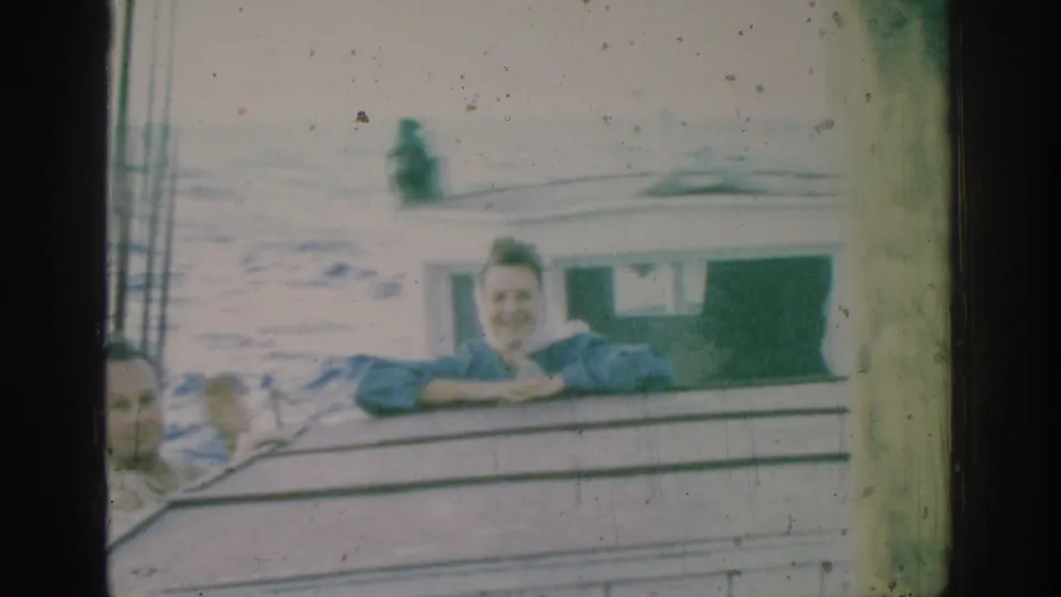 The woman in blue smiles from the boat rail during the 1962 Rhodes home-movie boat ride, with water visible behind her.