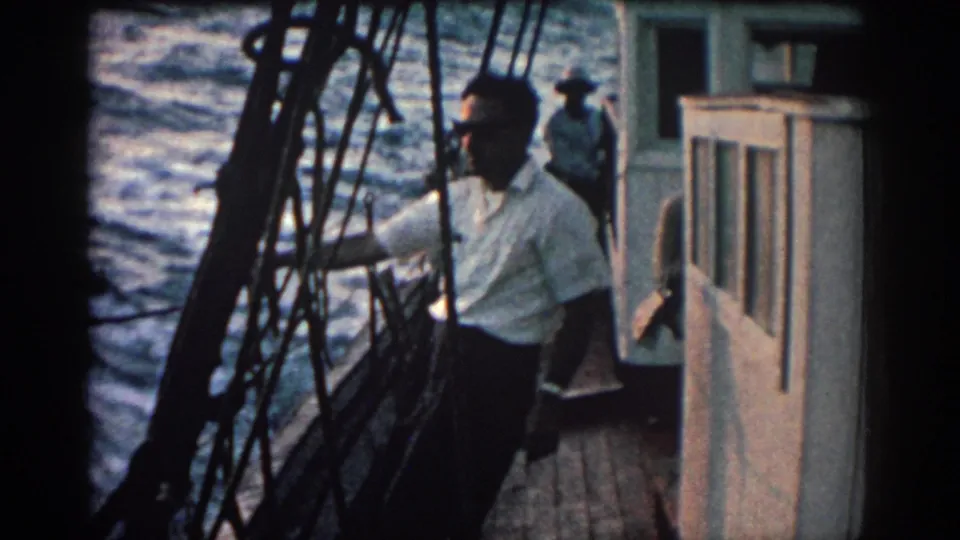 In 1962 Rhodes, Greece footage, a man stands on a boat deck near rigging and cabin windows as the sea moves behind him.