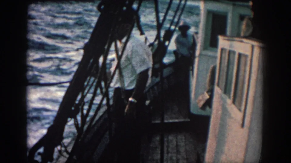 A man in a white shirt stands turned away on the deck while another person is visible near the cabin in 1962 Rhodes, Greece footage.