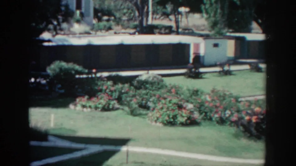 1962 Rhodes 8mm frame showing a wider landscaped garden with red flowers, trimmed lawn, white path, and a low building.