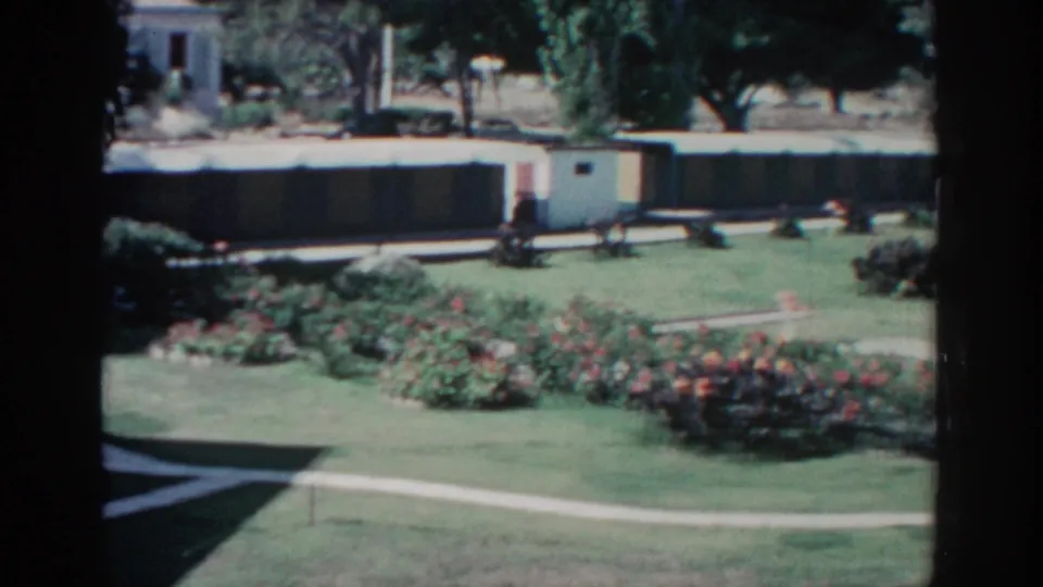 1962 Rhodes garden frame with a white path crossing the lawn, red flowering beds, shrubs, trees, and a low building behind.