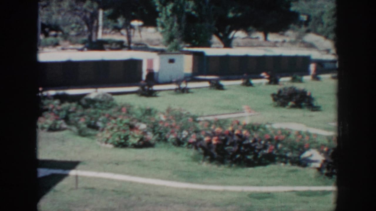Landscaped lawn with flowerbeds, curved paths, trees, and a long low building in the background.