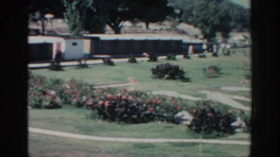 Rhodes, Greece garden scene from 1962 with flowering beds, walkways, trees, a low building, and distant walkers.