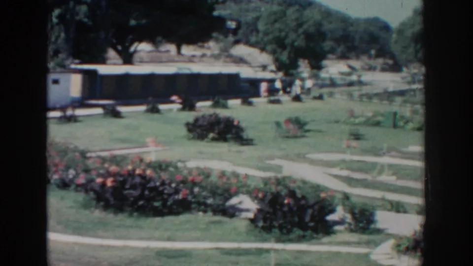 1962 home-movie frame of a Rhodes garden with curving paths, flowering borders, trees, and people walking in the distance.