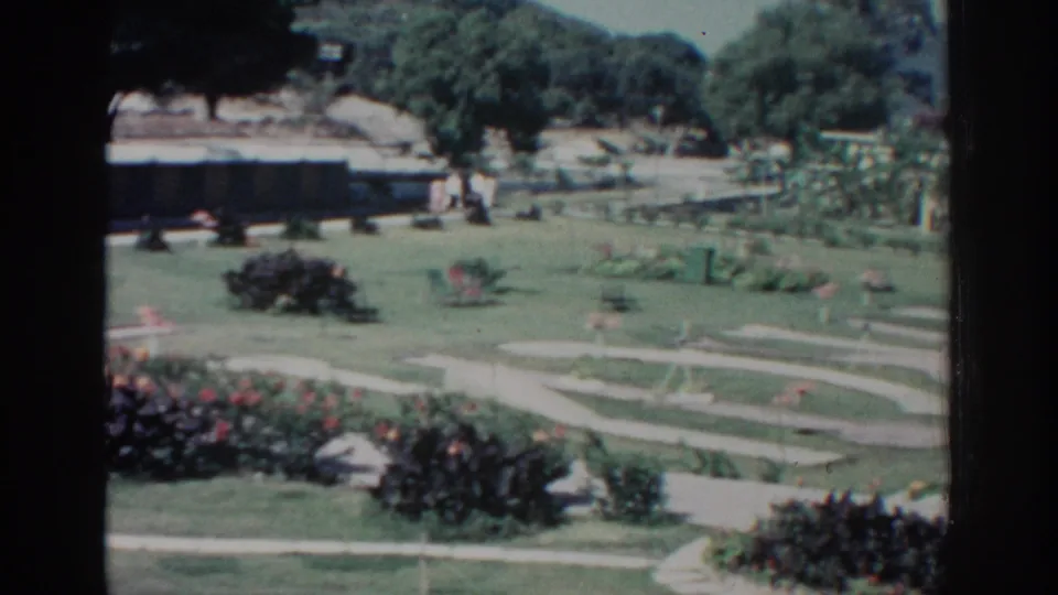 Rhodes garden in 1962 with intersecting walkways, planted beds, open grass, trees, and small distant figures walking.