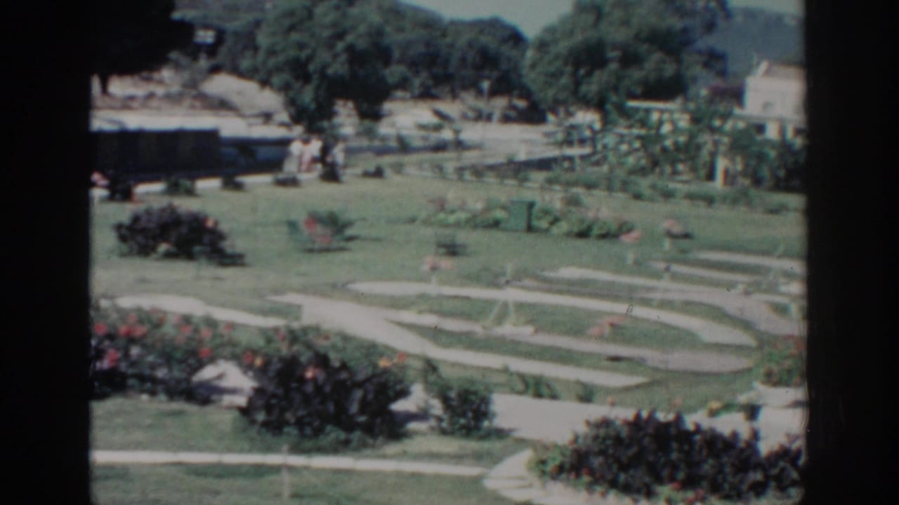 Sunlit garden with winding paths, lawns, flowerbeds, trees, and pale buildings in the distance.