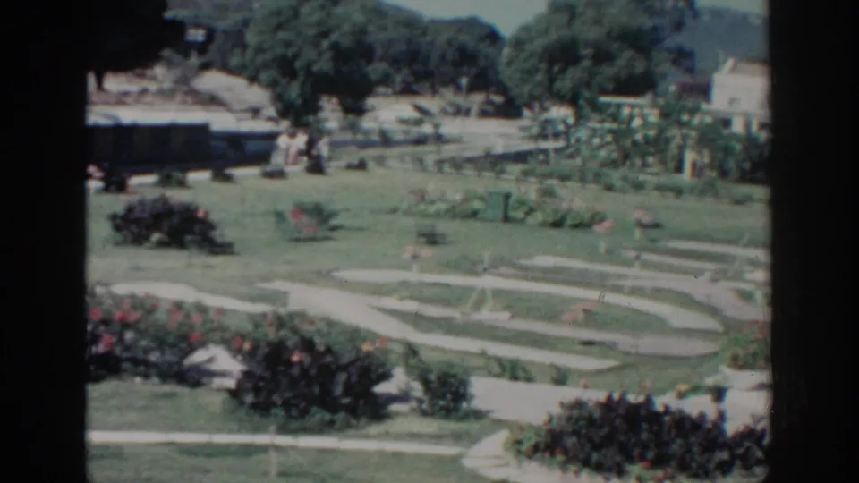 1962 8mm view in Rhodes, Greece, showing a landscaped garden with curving paths, red flowers, trees, distant hills, and pale buildings.
