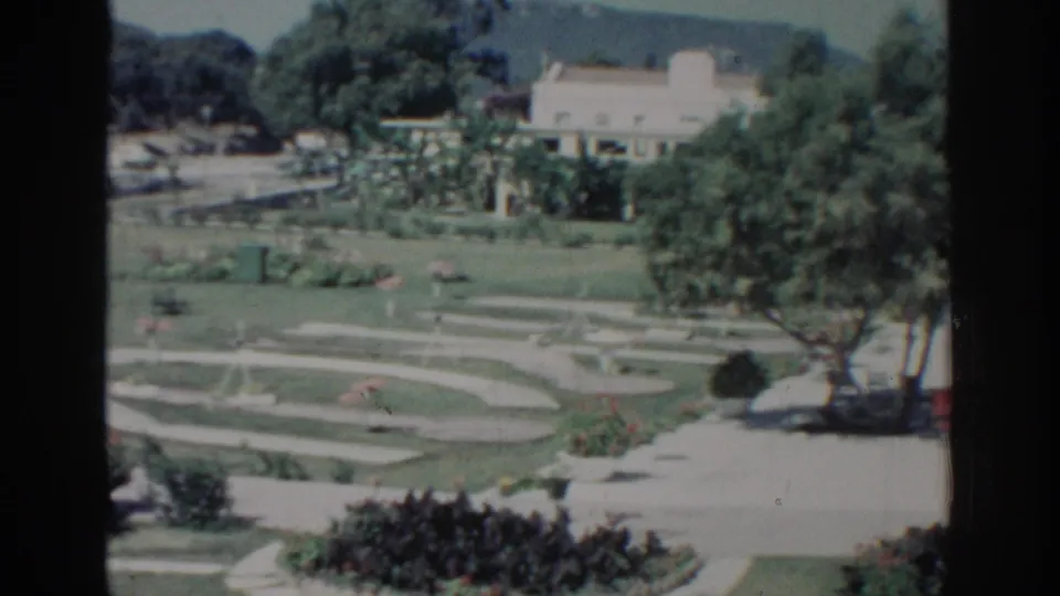Rhodes, Greece garden footage from 1962 with a pale building behind trees, winding paths, green lawns, red plantings, and distant hills.