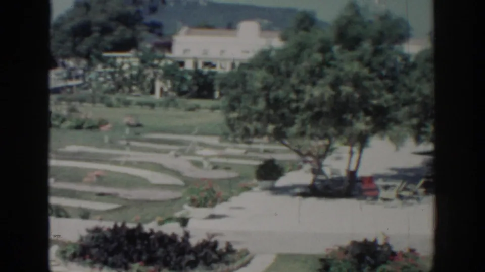 Late frame from 1962 Rhodes 8mm footage showing curving garden paths, lawns, red flowers, shade trees, benches, and a pale building.