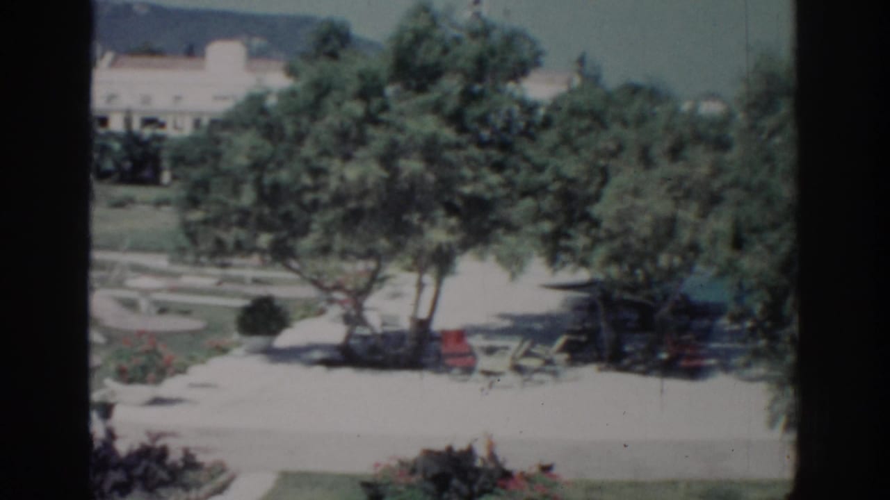 Rhodes, Greece garden scene from 1962 with shaded trees, bright paving, outdoor seating, lawn beds, and a distant light-colored building.