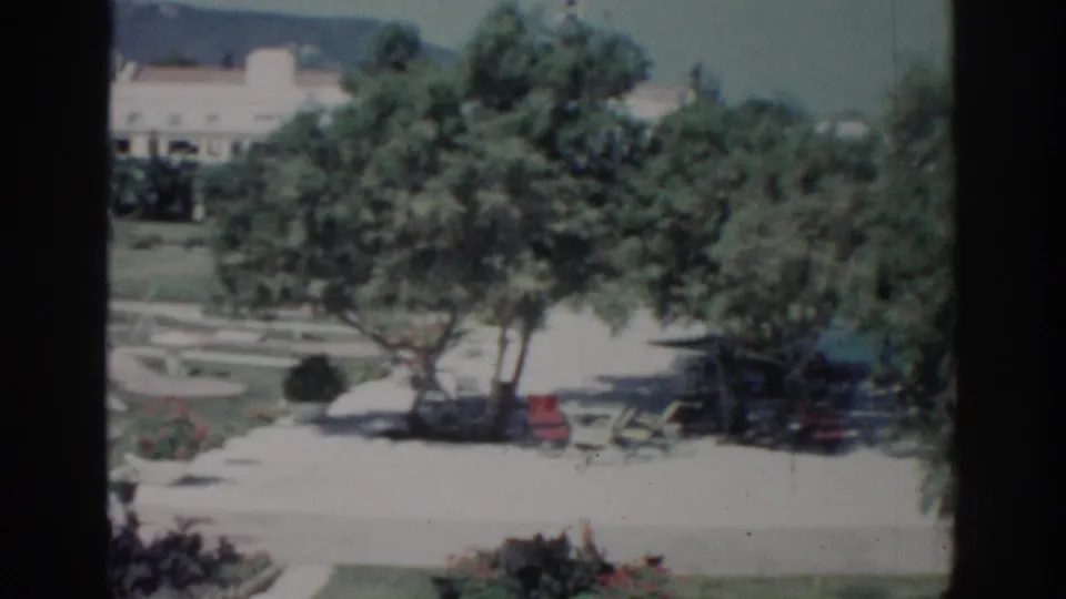 Rhodes, Greece garden scene from 1962 with shaded trees, bright paving, outdoor seating, lawn beds, and a distant light-colored building.