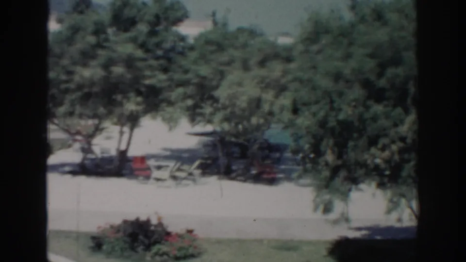 1962 Rhodes home-movie frame showing shaded garden seating, a paved path, lawn, flower beds, and leafy trees in bright sun.