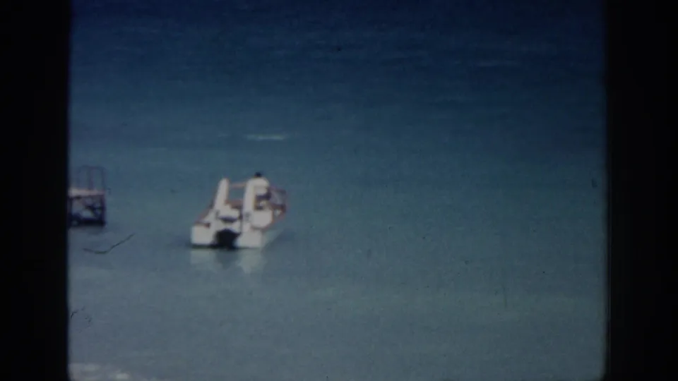 1962 Rhodes coastal frame showing a white motorboat on calm turquoise sea, with a small dock at the left edge.