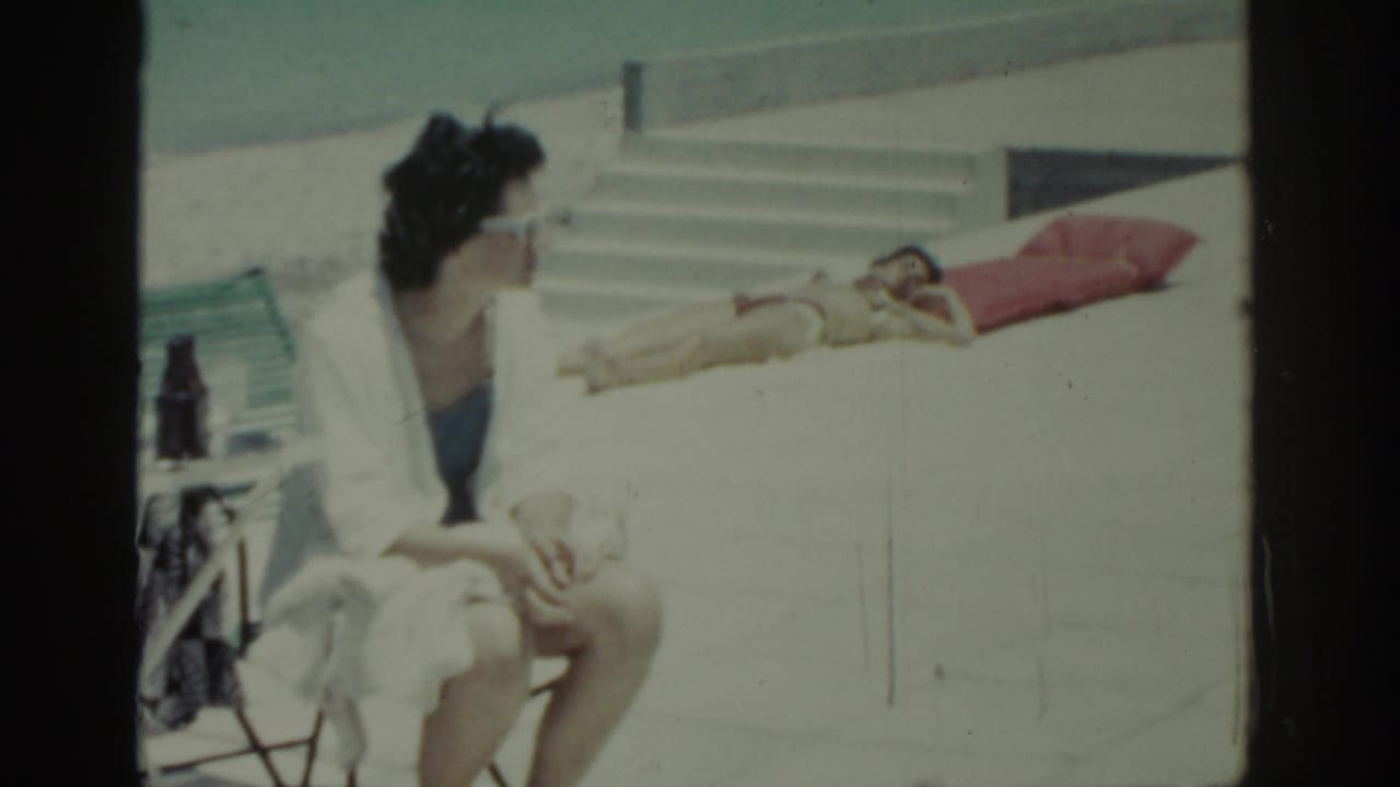 1962 Rhodes resort scene with a seated woman in a white robe and blue swimwear facing a sunbather on a red towel near steps.
