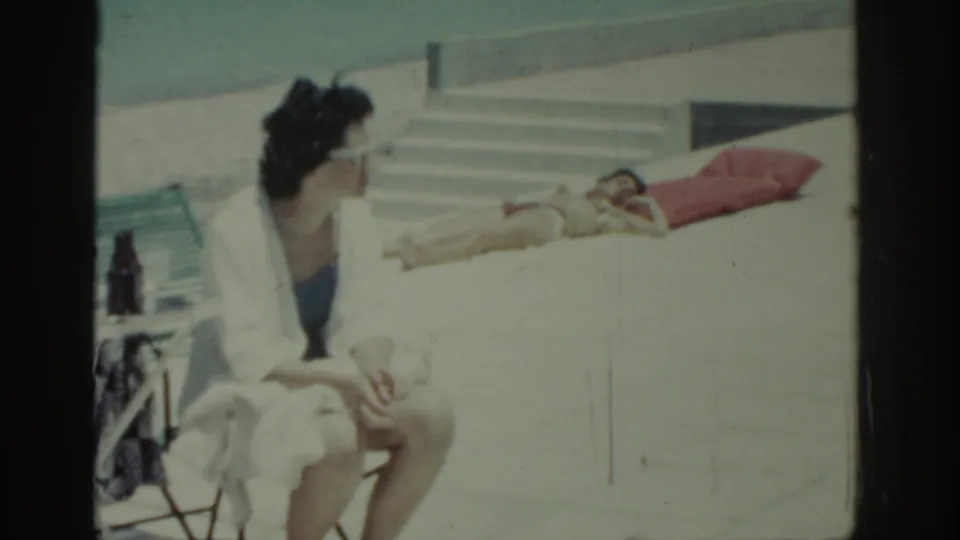 1962 Rhodes resort scene with a seated woman in a white robe and blue swimwear facing a sunbather on a red towel near steps.