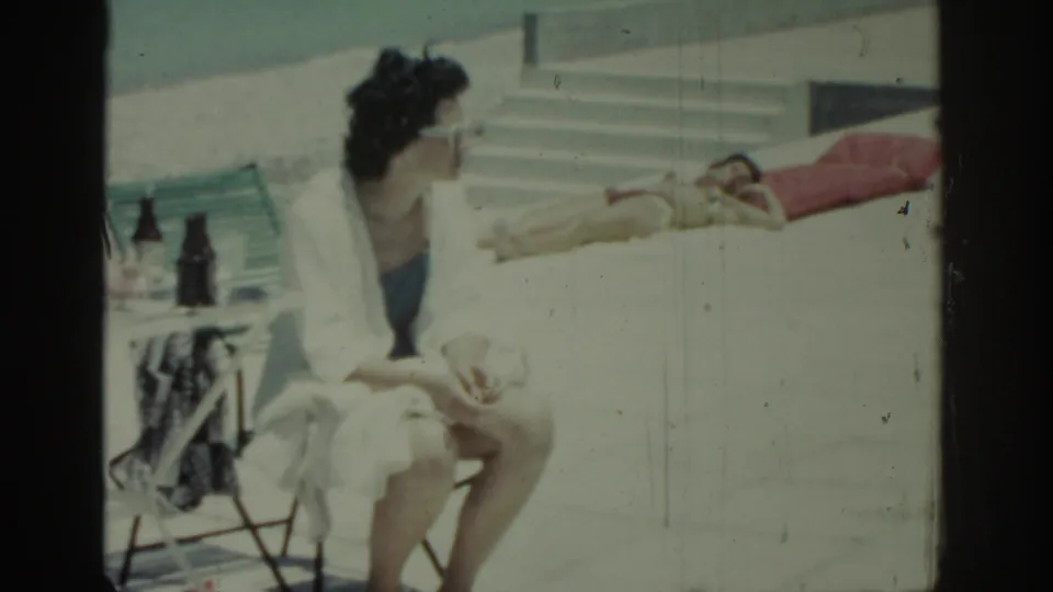 Rhodes, Greece home-movie frame showing a woman in a white robe seated near patio furniture and a sunbather on a red towel.