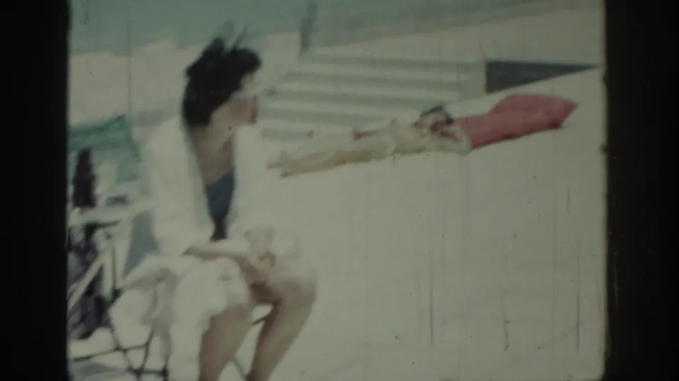 Vintage 8mm frame from Rhodes showing a seated woman in white over blue swimwear, with a reclining sunbather in the background.