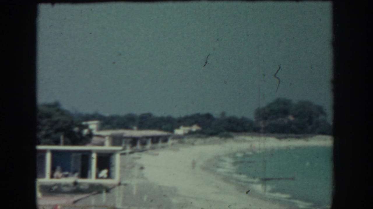 1962 Rhodes home-movie frame showing a pale sandy shore, calm turquoise water, low waterfront structures, trees, and tiny beachgoers.