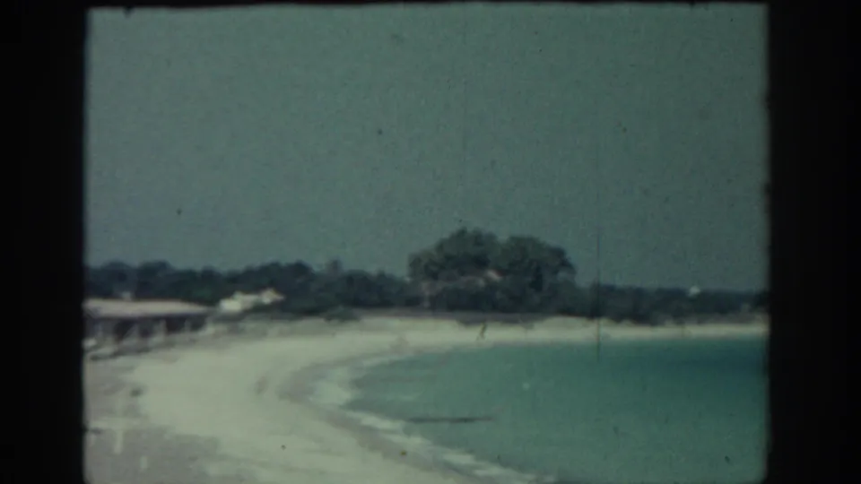 1962 Rhodes coastal frame with a curving sandy beach, quiet blue-green water, low shore structures, and trees beyond the sand.
