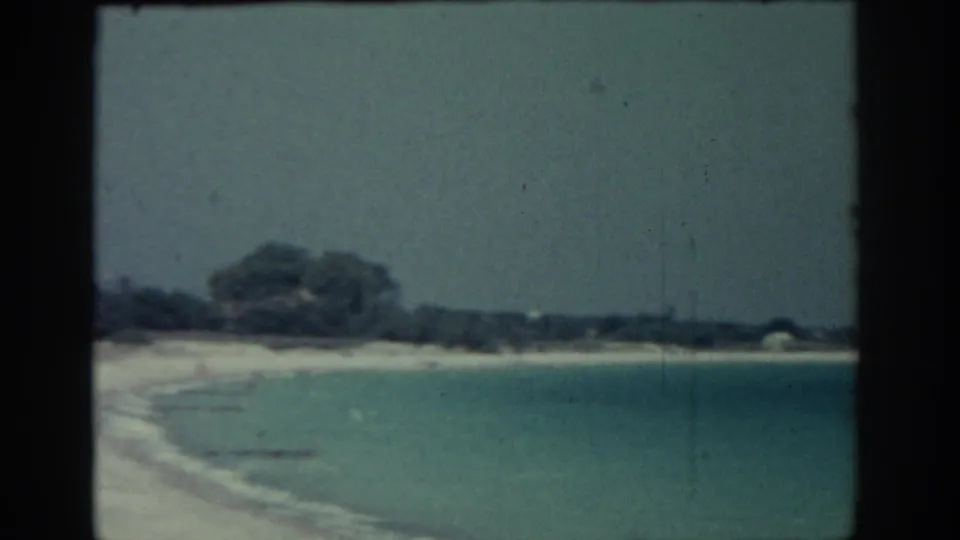1962 Rhodes home-movie view of shallow coastal water beside a sandy beach, with trees and a small pale structure in the distance.