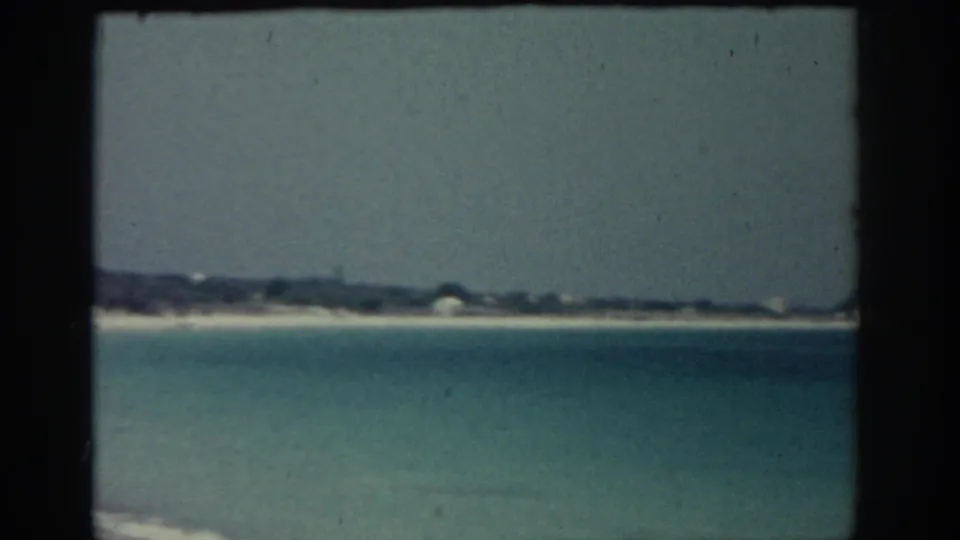 1962 Rhodes coastal frame looking over calm blue-green water toward a sandy shoreline and low distant features.