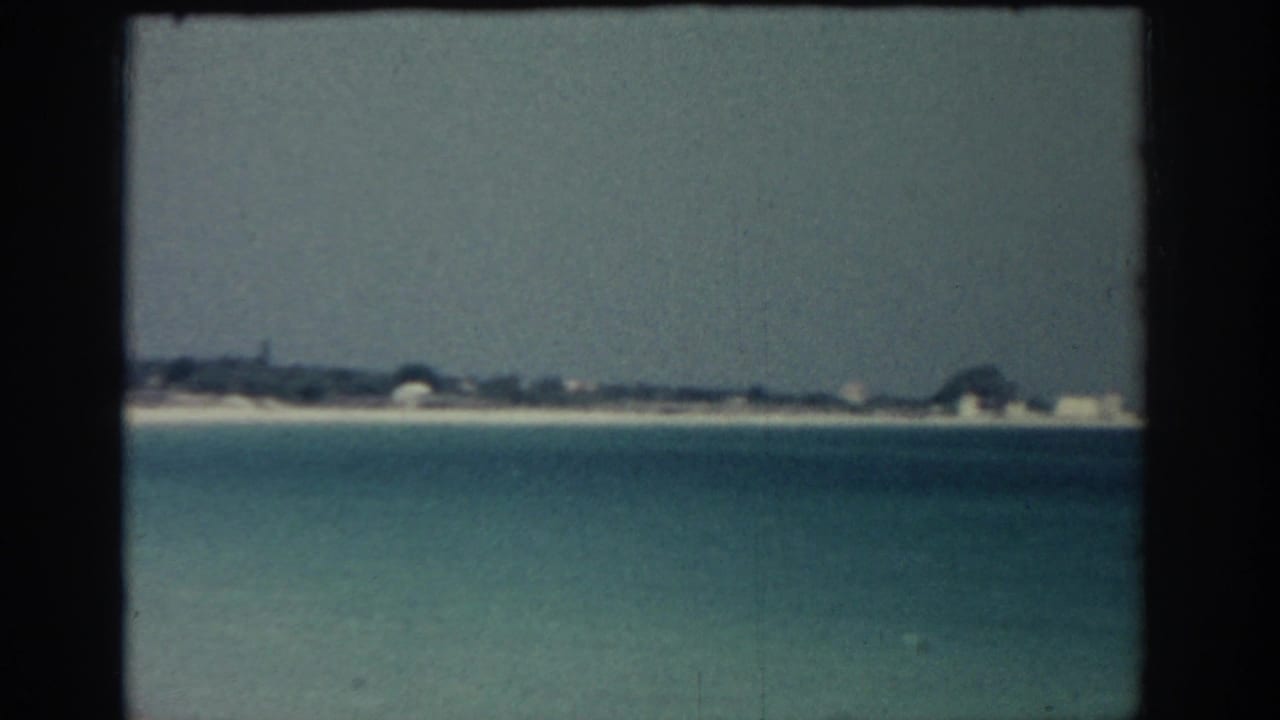 Turquoise sea facing a pale beach and distant low buildings under a hazy sky.