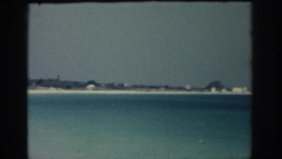 1962 8mm view from Rhodes, Greece, looking across blue coastal water toward a sandy shoreline with low buildings.