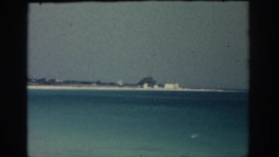 Rhodes coastline in 1962, with calm blue water in the foreground and pale beach structures along the horizon.