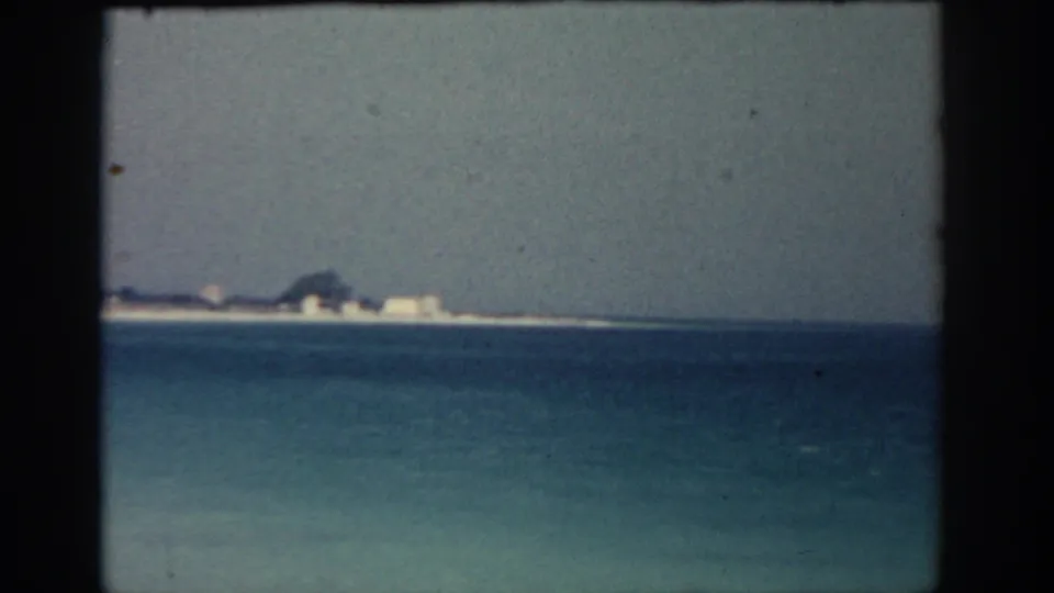 Soft 8mm frame of the Rhodes shore, showing blue water, a pale beach line, trees, and low white coastal structures.