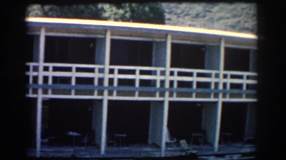 1962 Rhodes waterfront building with white railings, open shaded bays, and trees behind it, filmed in soft 8mm color.